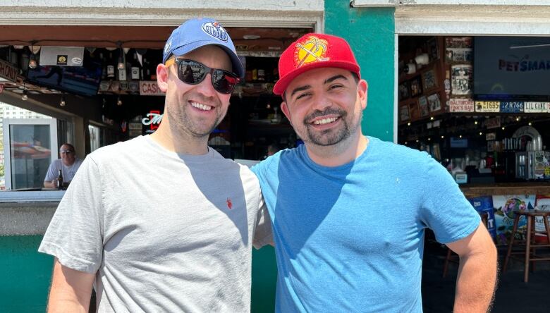 Two men are standing in the sunshine in front of a bar. The man on the left is wearing a grey t-shirt and blue Edmonton Oilers ball cap. The man on the right is wearing a blue t-shirt and a red Florida Panthers hat.