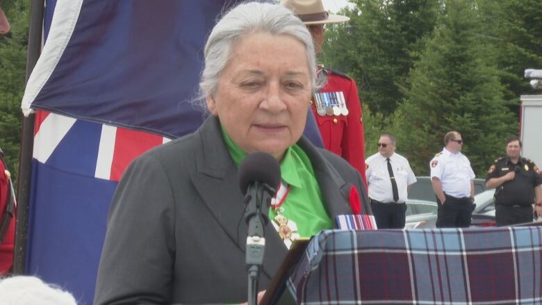 A woman with white hair stands at a microphone outside with people behind her.