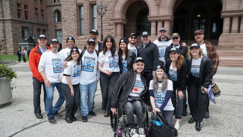 A group of people in ACTRA shirts pose before the staircase of a large old brick building. 