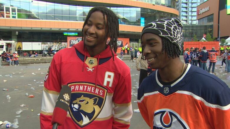 Two black men, one in a red hockey jersey and the other in an orange-and-blue jersey, are standing outside in downtown Edmonton, while speaking to someone holding a microphone.