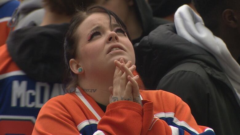 A white woman, with dark hair pulled back in a pony tail, is clasping her hands together. She is wearing a blue-and-orange hockey jersey, while outside among a crowd.