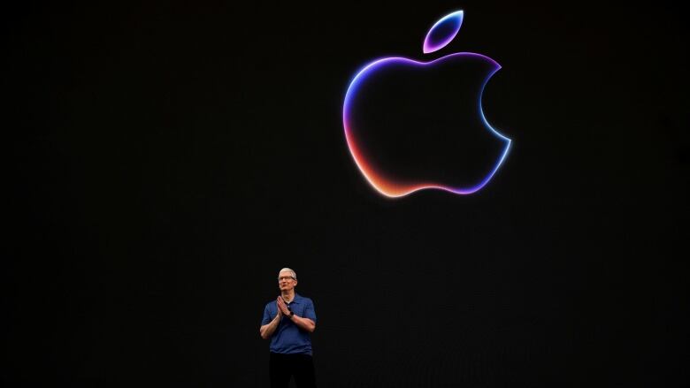 A man stands on a dark stage with a large multicoloured illustration of an apple glowing behind him.