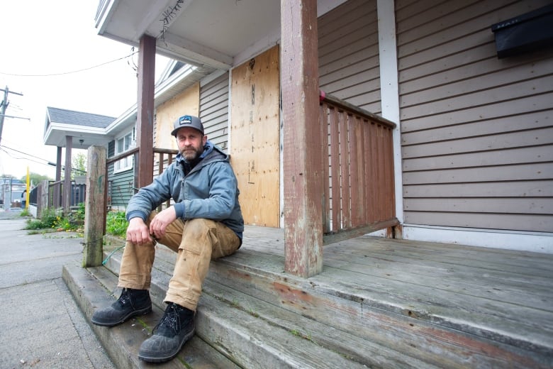 A man wearing a jacket, hat and work boots sits on the step of a boarded up residence.