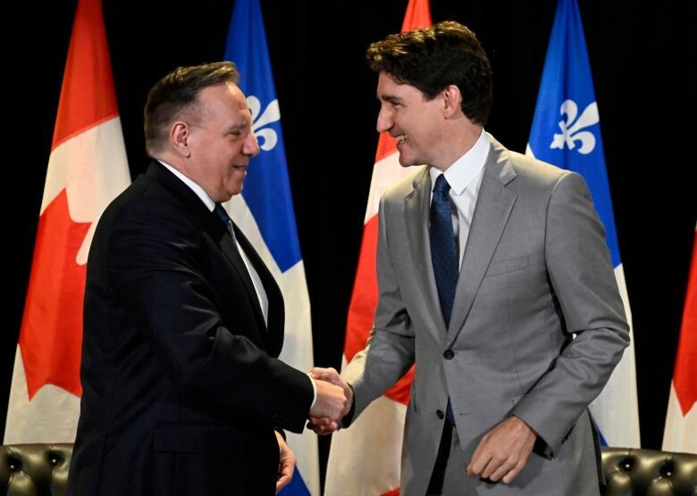 Two men shake hands, with Canadian and Quebec flags behind them.