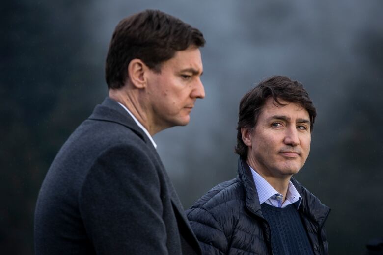 Two tall men wearing formal clothes are pictured outside. One is looking down towards the ground, while the other looks at the camera.