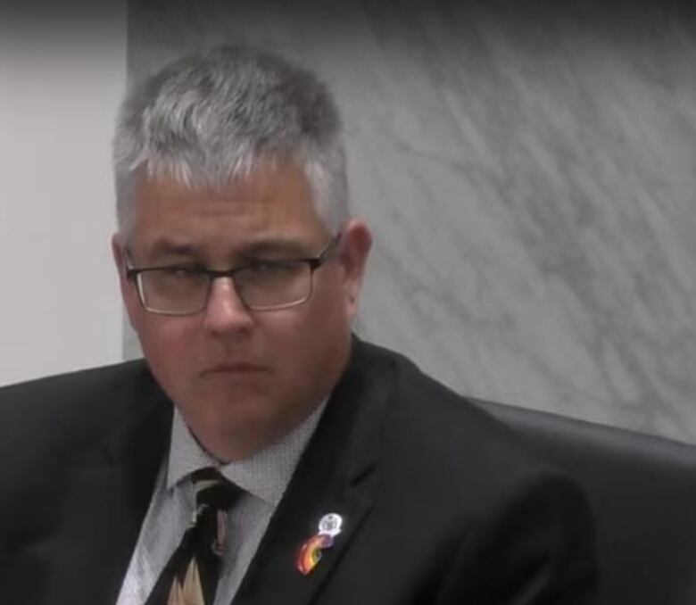 Man with gray hair sitting at a table with mic in front of him with neutral expression.