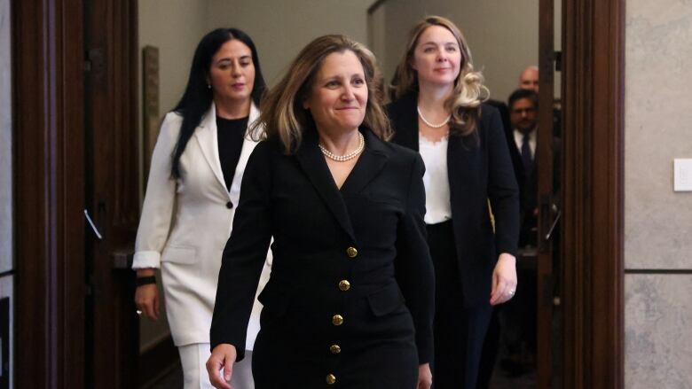 Three women in business suits walk through a set of double door. The woman in the middle front, Chrystia Freeland, wears a set of pearls and a confident smile.