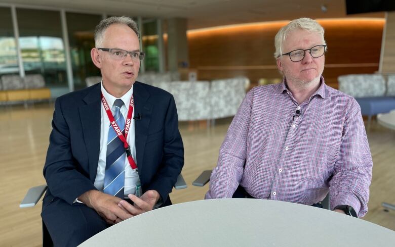 Man with grey hair, a blue suit and glasses sits next to a man with white hair and glasses wearing a purple shirt.  