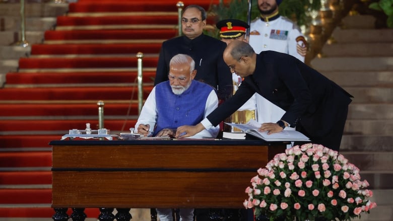 A man in a white shirt and blue vest sits at a desk and signs documents. A man dressed in black points at the document, while another stands behind him. At the back is a man in white. 