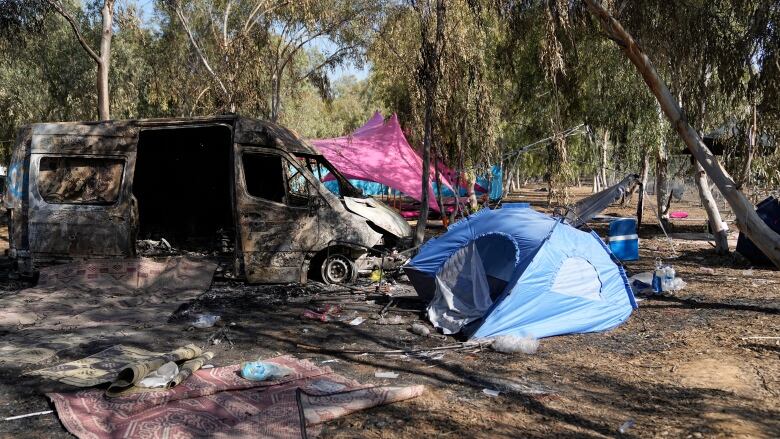 A burned-out vehicle and several tents are shown in a rural-like setting with several trees shown.