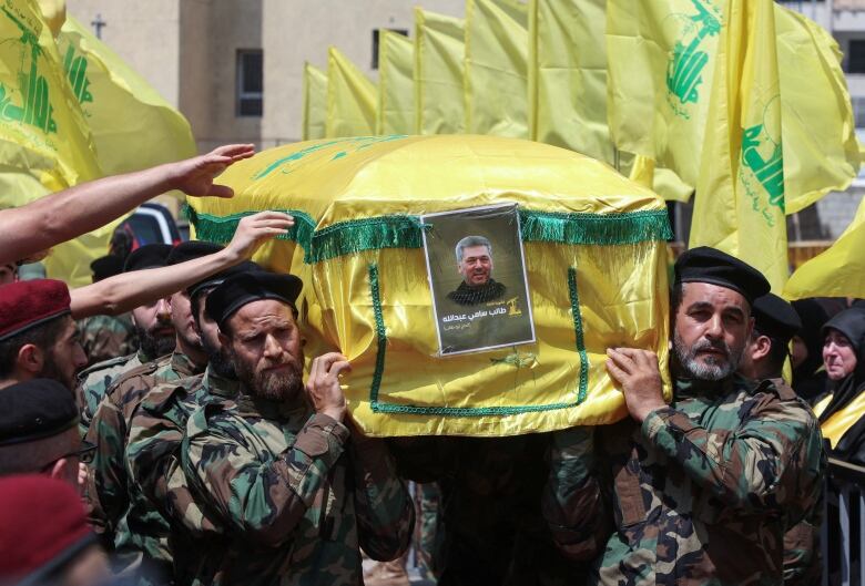 Several men in military fatigues and caps carry a coffin draped with multicoloured cover.