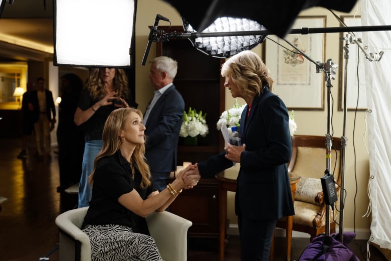 A seated woman shakes hands with a standing woman. Behind them are lighting fixtures and camera equipment. 