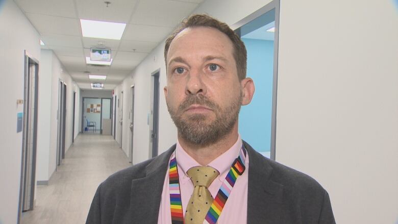 A man standing in a hallway looking off-camera. He is wearing a pink shirt, gold tie, grey suit jacket, with a rainbow lanyard around his neck.