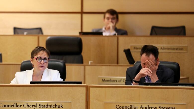 A woman in a white suit and a man in a black suit sit at a wooden desk. The man has his head in his hands. 