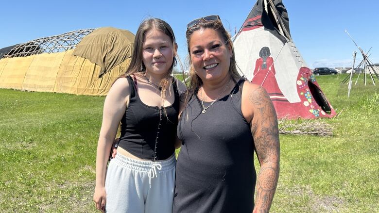 Two women smile together in a field in front of a teepee.