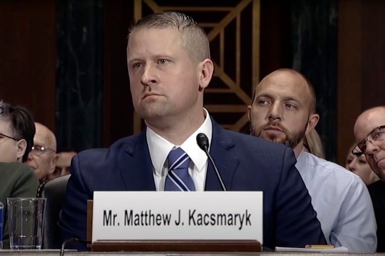 A man with short hair, clean shaven and wearing a suit and tie, is shown seated at a table at a hearing.