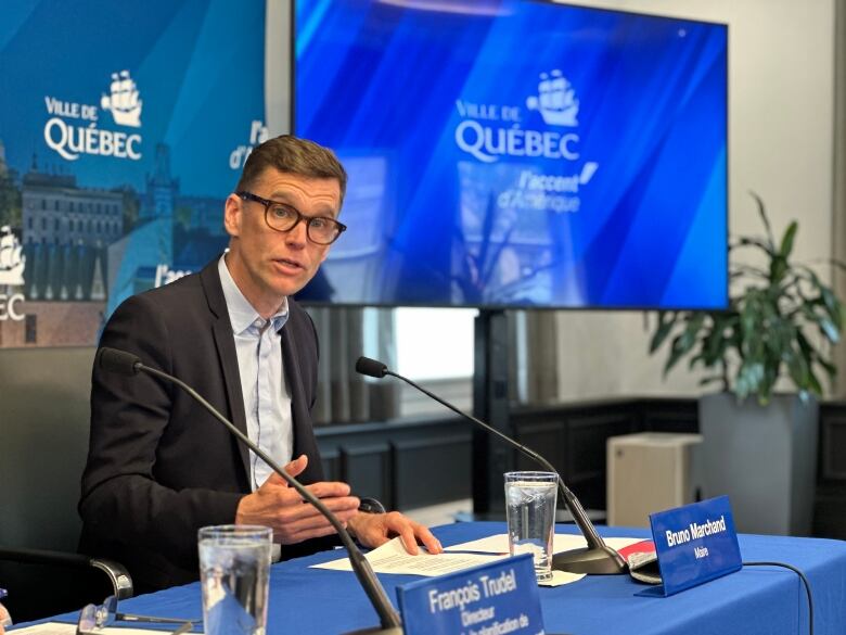 A man in a suit speaks sitting at a table, in front of a microphone