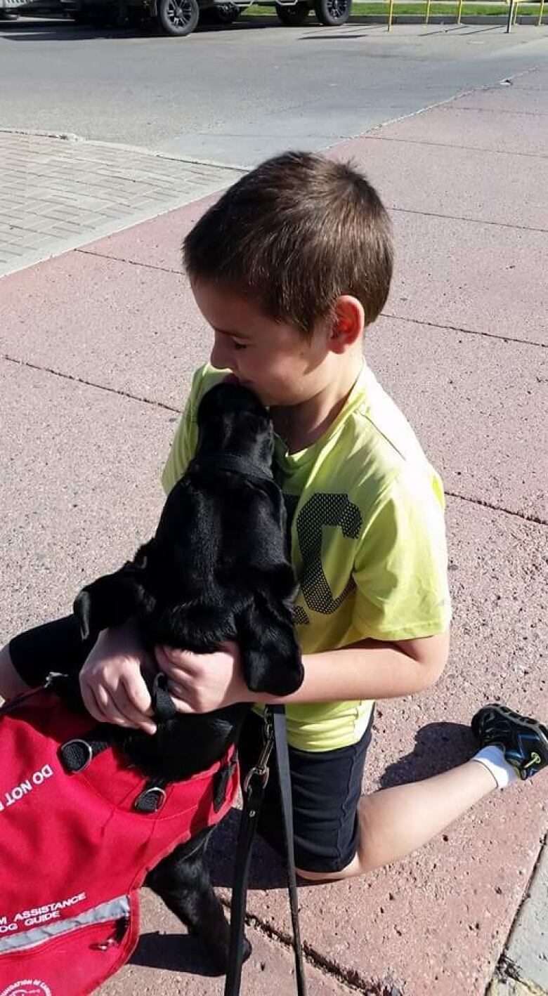 A black dog in a red vest licks the face of a boy in a yellow shirt.