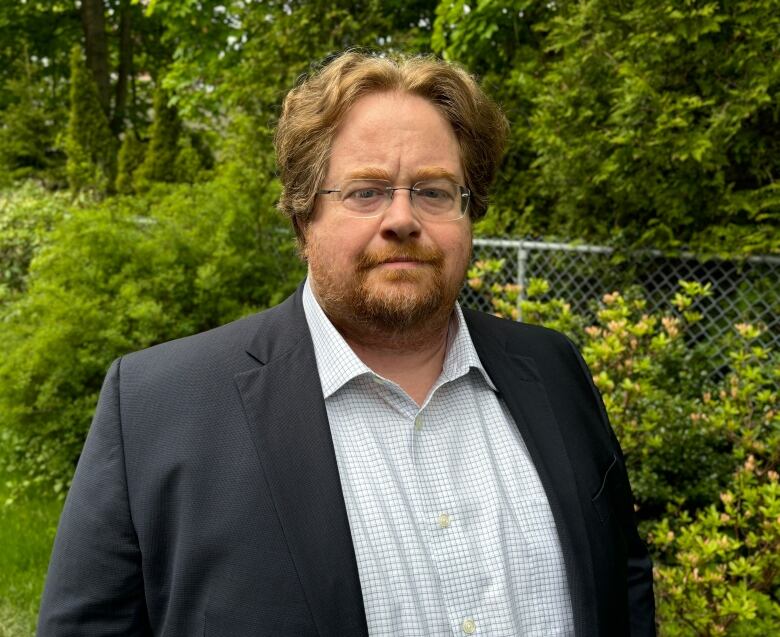 A person wearing a dress shirt and suit jacket stands outside in front of greenery.