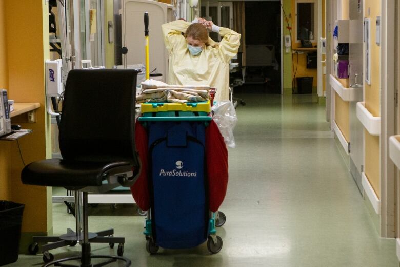 A woman wearing a surgical mask reaches behind her head to tie a personal protective gown while standing behind a cart filled with cleaning supplies in a hospital hallway. 