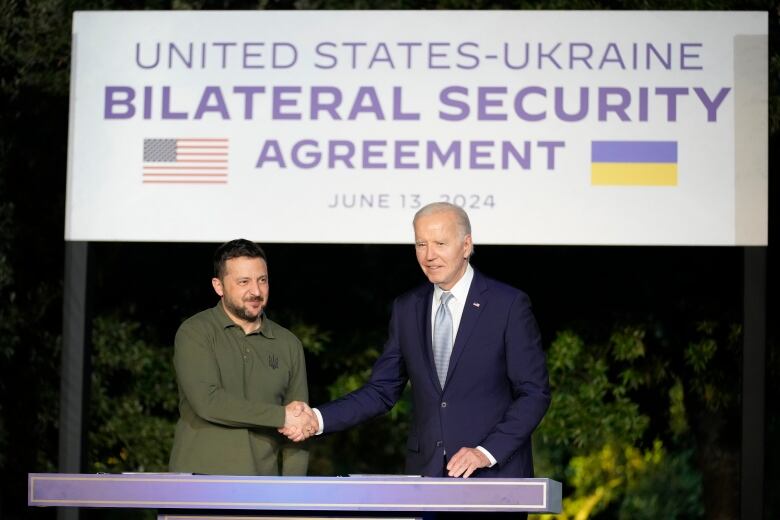 A bearded man in military fatigues shakes hands with an older clean shaven man wearing a suit and tie as they stand behind a podium.