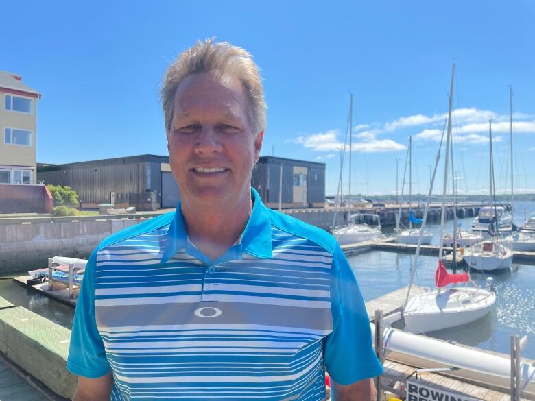 A blonde man stands on the marina smiling for a photo. His shirt is blue with white stripes. There are sailboats behind him.