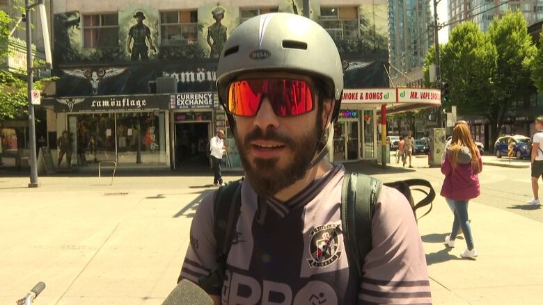 A man wearing a motorcycle helmet and large sunglasses speaks on a city street.