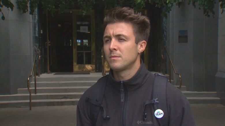 A white man wearing a zip-up jacket and a white pin badge speaks outside a city hall.