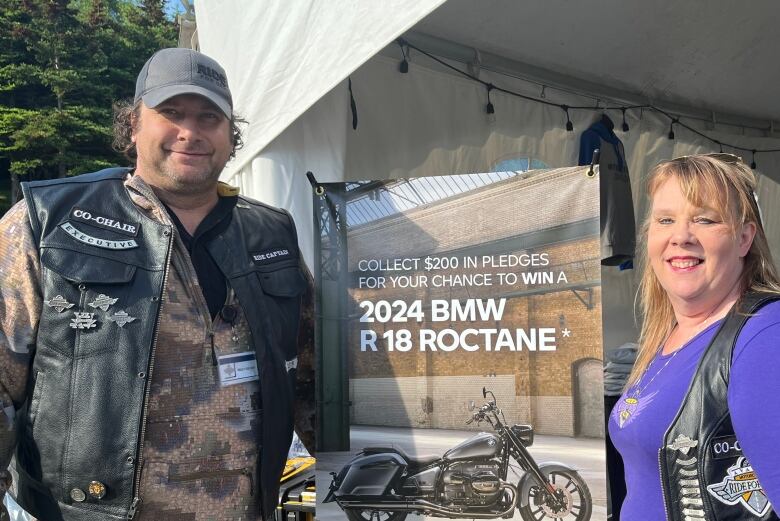 A man, on the left, and a woman, on the right, smile. They both wear motorcycle vests. In between them, there's a fundraising poster.