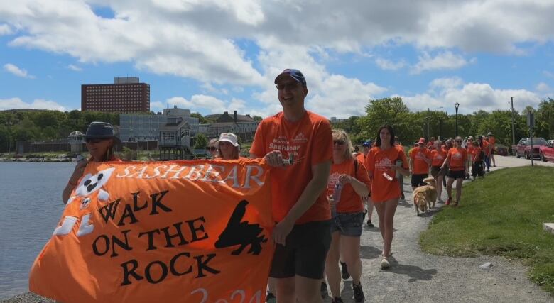 A large crowd of people wearing orange shirts are seen alongside a body of water. Two people nearest the camera are holding a banner that reads 