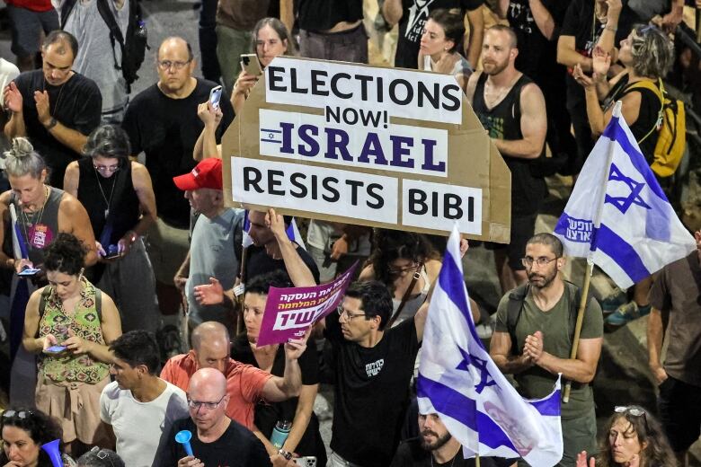 A crowd shot is shown, with dozens of people shown standing and chanting at a protest. One held up a sign that says, 'Elections Now - Israel resists Bibi.'