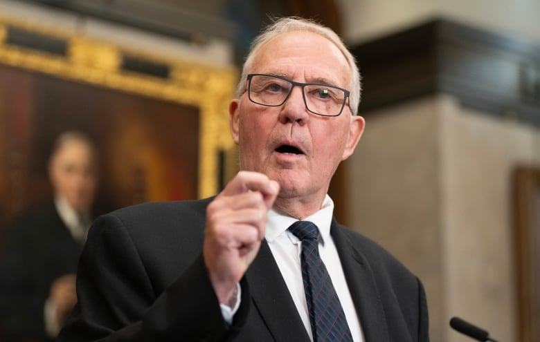 A man in a suit with Grey Hair holds his hand up as he speaks to reporters from the foyer of the House of Commons. 