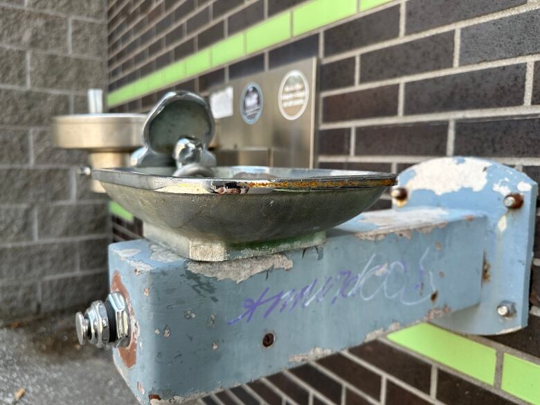 A rusty drinking fountain is shown with graffiti on the side.