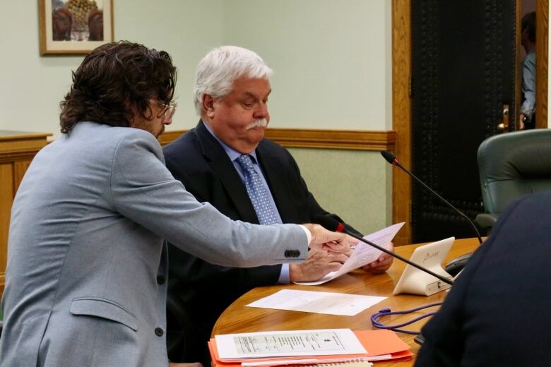 A man in a light blue suit points to a piece of paper being held by a man in a black suit. Both are sitting at a wooden table. 