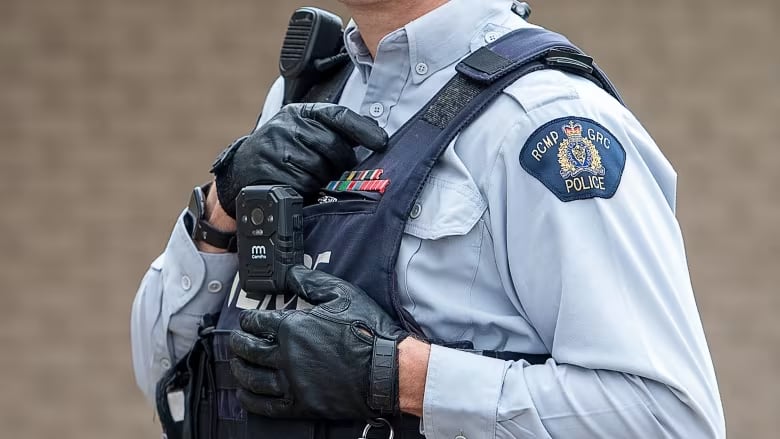 A Mountie wearing a grey uniform and leather gloves attaches a body worn camera to their bullet proof vest. 