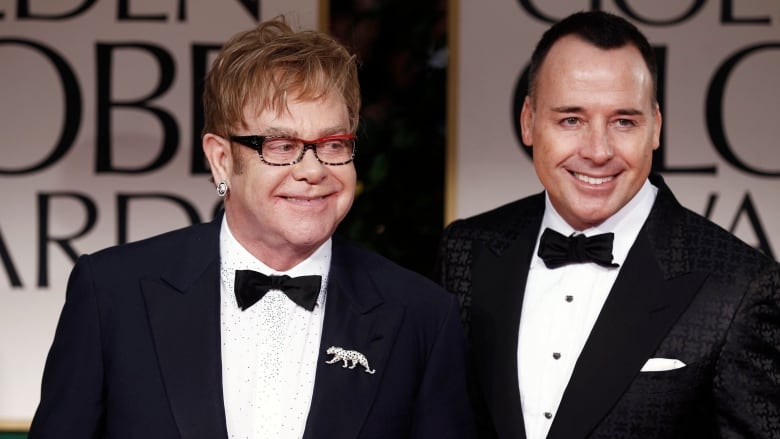 Two smiling men wearing tuxedos pose.