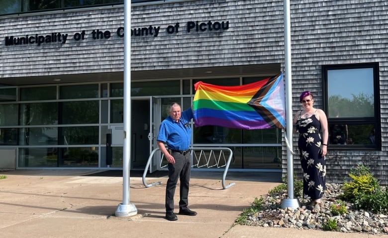 Two people stand by a flag pole, and hold up a Pride flag.