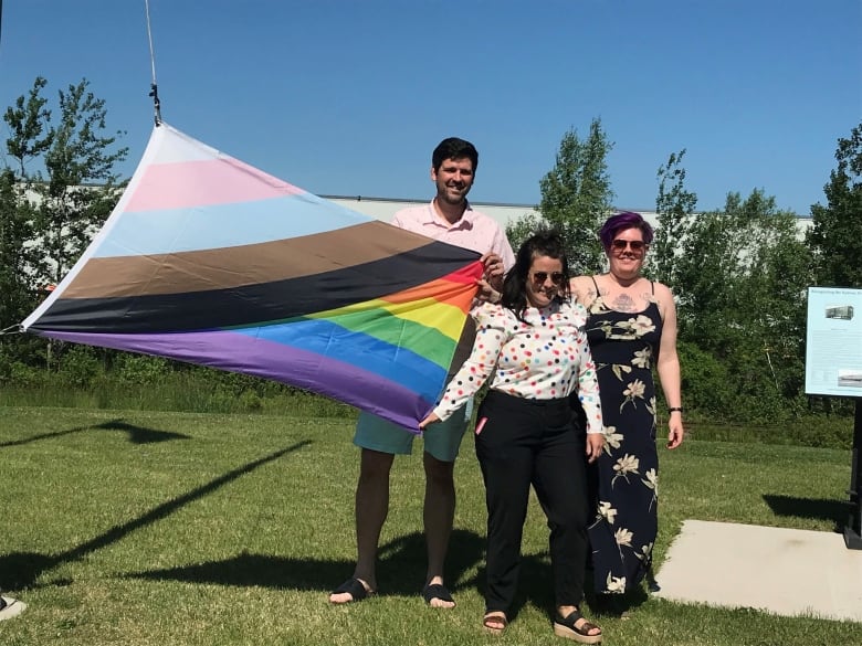 Three people stand next to a large Pride flag and smile for a photo.