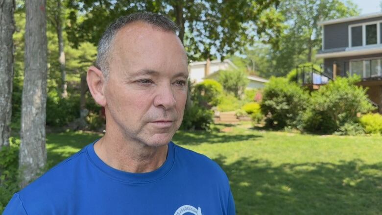 A man in a blue t-shirt stands on a lawn while speaking to the camera.