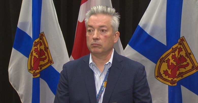 A man with white hair wears a suit jacket and white striped shirt. He stands in front of two Nova Scotia flags with a Canadian flag in the middle. 