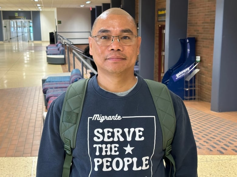 A man with a shaved head and glasses poses in a university building hallway. He is wearing a backpack and a blue sweatshirt that says 