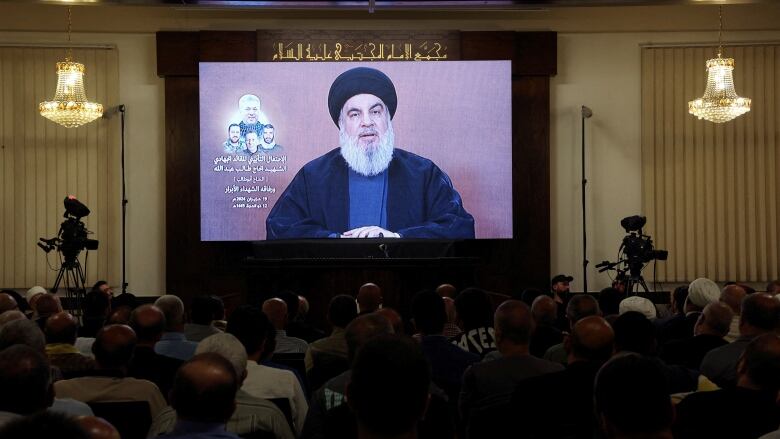 An older bearded man with a religious head covering is shown projected on a screen as dozens of people are shown seated in a room watching and listening.