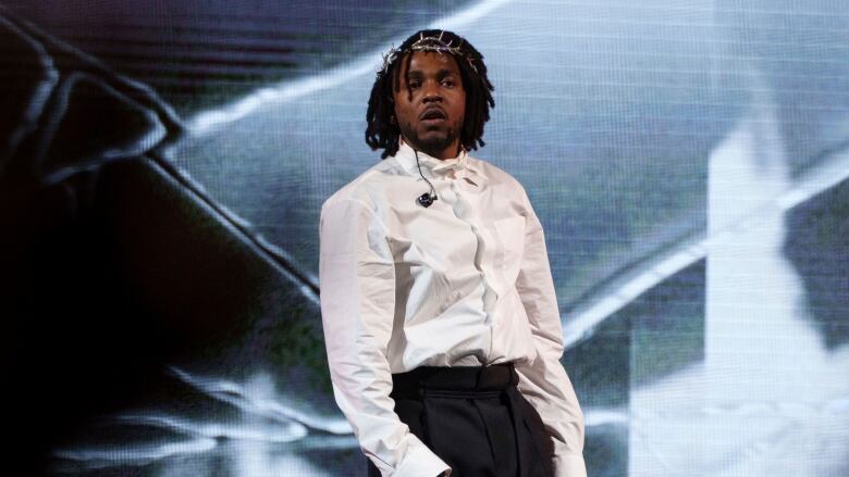 A man in a crumpled white dress shirt, black pants and a silver barbed wire crown on his head holds a microphone performs on stage, with projections seen behind him.