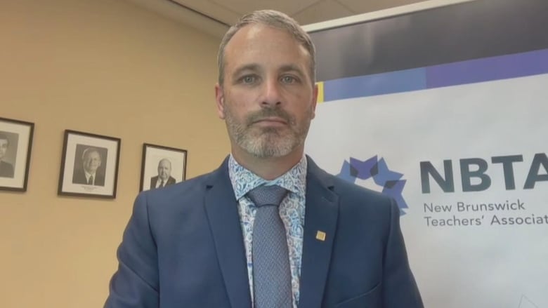 A person in a suit and tie stands in front of a New Brunswick Teachers' Association banner.