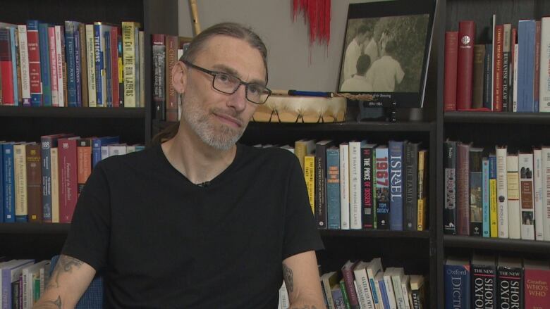 A man in a black t-shirt sits in front of a bookshelf.