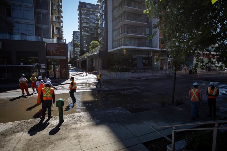 Men in orange and yellow vests stand in dirty water in the middle of a road.