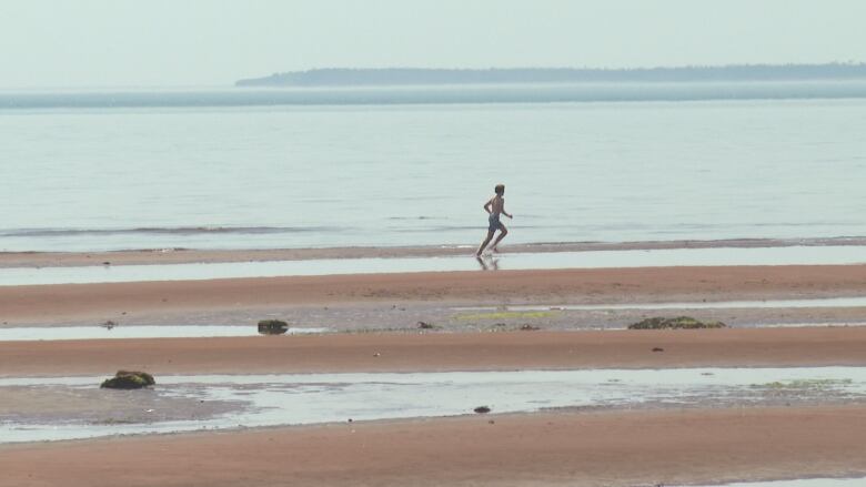 A man in swimming trunks runs across a red sand beach with water in the background.