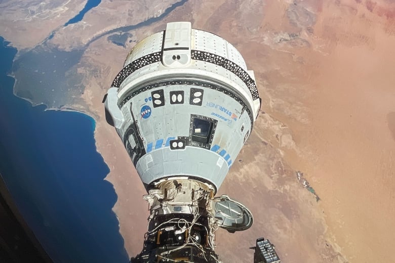 Earth is in the background with the ocean and desert, and in the foreground is a white spacecraft attached to the International Space Station.