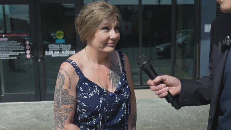 A woman wearing a blue top with tattoos on her shoulder speaks to a person with a mic.