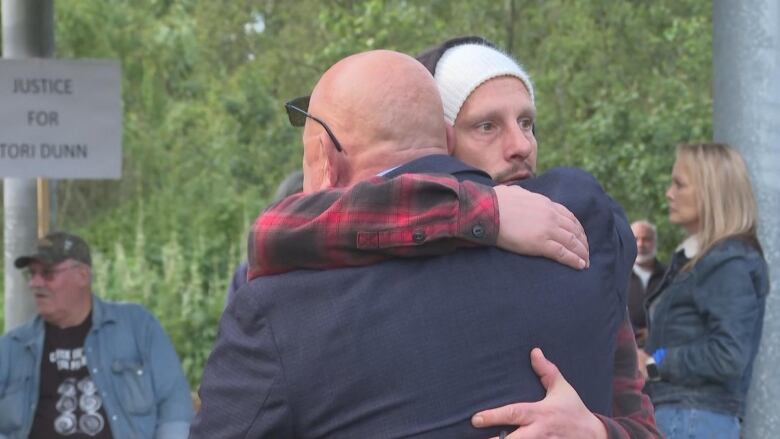 A bald man and a man in a beanie hug in an outdoor environment.